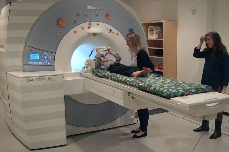 MIT graduate student Hilary Richardson helps a child into an MRI scanner for a study of how children's brains develop the ability to think about the mental states of other people.Richardson shows the brain scans to one of the children participating in the study.