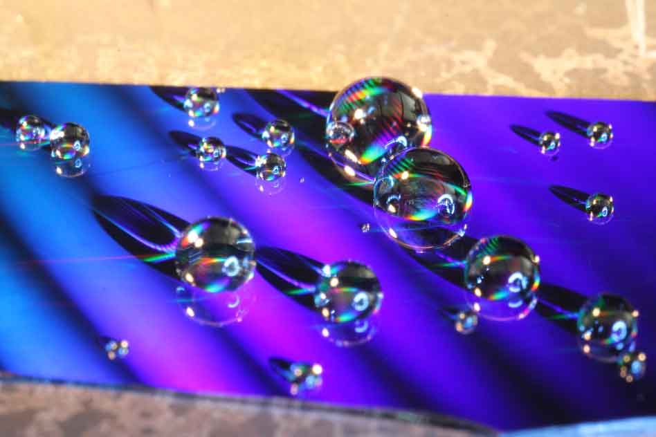 This photo shows water droplets placed on the nanostructured surface developed by MIT researchers. The colors are caused by diffraction of visible light from the tiny structures on the surface, ridges with a specially designed shape. Images: Kyle Wilke