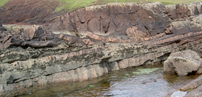 A field photo take at Stoer showing the laminar beds of sandstone in the bottom of the picture. In the middle is the impact deposit (12m thick at this location) that contains 