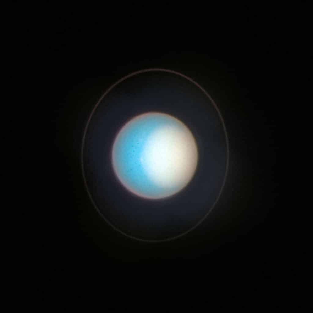 Uranus appears tipped on its side. Set against a black background, the planet is mainly colored cyan. It looks like a flat circle outlined with a pinkish gray limb. A faint, pink ring encircles the planet nearly vertically. The faint ring appears to be almost face on. A large area of white coves much of the right side of the planet.