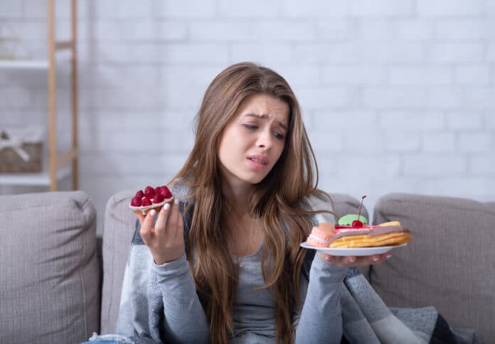 Depressed young lady having eating disorder, comforting herself with sweets on sofa at home. Unhappy millennial woman coping with negative emotions through food, suffering from bulimia
