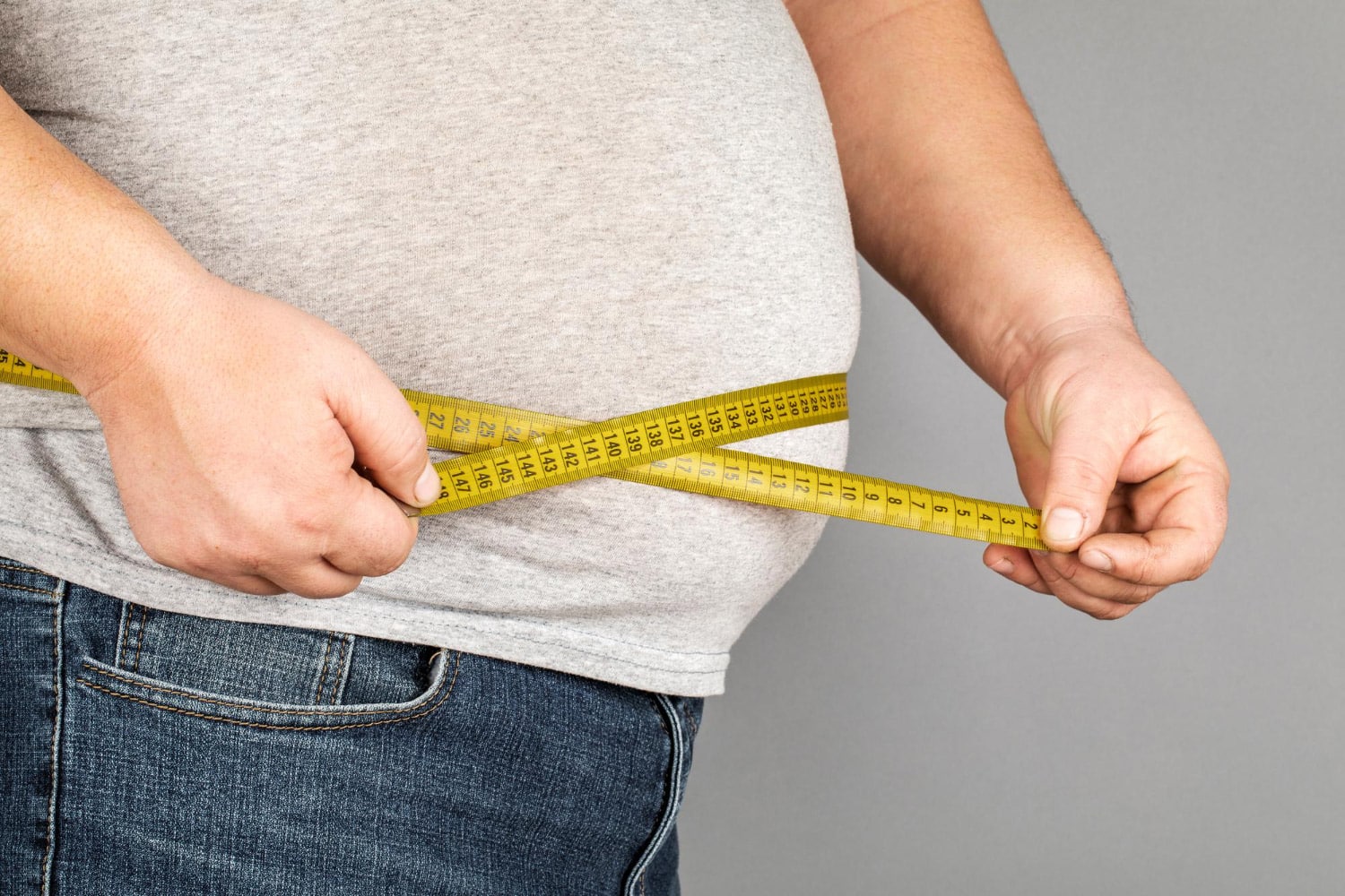 Image showing a man measures his fat belly with a measuring tape on a gray background.