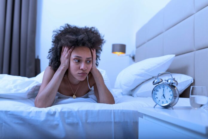 Sleepless and desperate woman awake at night not able to sleep, feeling frustrated and worried looking at clock suffering from insomnia in sleep disorder concept.