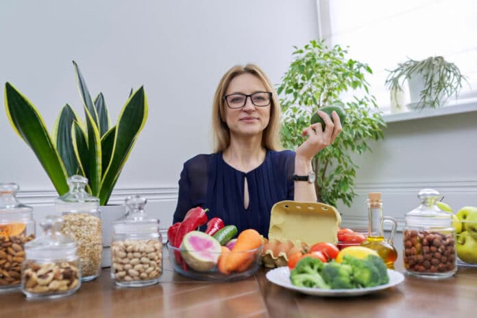 Middle aged woman professional nutritionist sitting at table with vegetables fruits eggs miasl nuts
