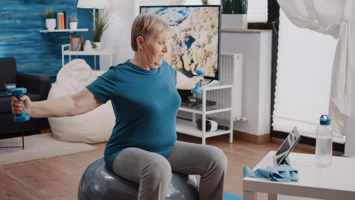 Aged woman watching video of workout lesson on tablet while doing physical exercise with dumbbells and sitting on toning ball. Old person following online training program with coach.