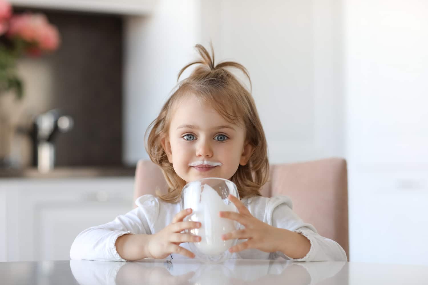 Blond cute child with blue eyes with traces of milk on the lips is holding a glass of milk siting at a white table in kitchen